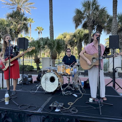 Three musicians perform outdoors on a stage with palm trees in the background. One plays bass, one drums, and one guitar.