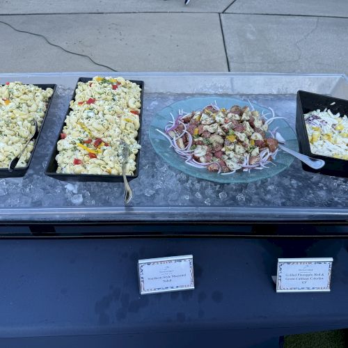 The image shows a buffet table with various salads, including pasta and potato salad, displayed in trays on ice.