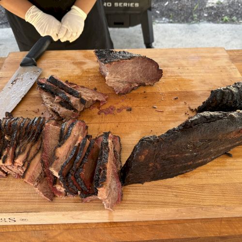 Slices of smoked brisket on a wooden cutting board with a large knife nearby. A person wearing gloves stands in the background.