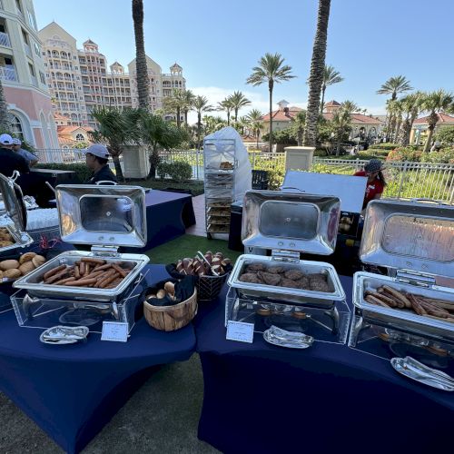 Outdoor buffet setup with covered food trays, various pastries, and dishes on tables. Palm trees and buildings in the background.