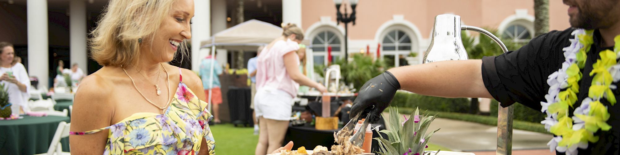 A woman in a floral dress receives food from a man at an outdoor event with a roasted pig on display.