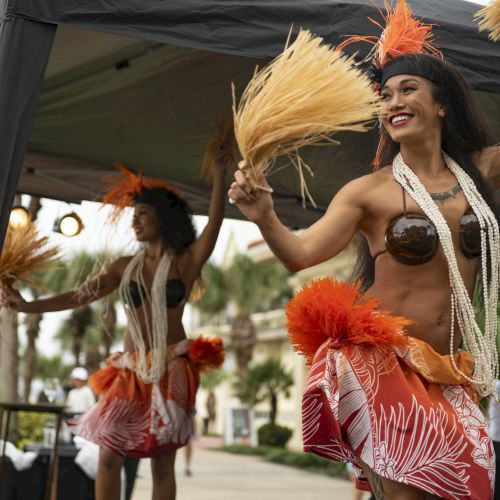 Two dancers in vibrant costumes perform energetically under a canopy, surrounded by palm trees, with an audience watching.