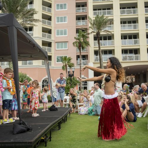 A hula dancer in a red skirt performs for a group of children on stage, with an audience seated on chairs outdoors in front of a hotel.