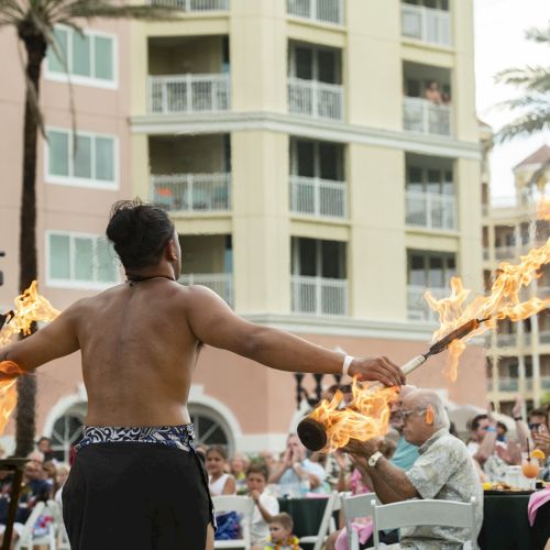 A performer is showcasing a fire dance with torches. The audience is outdoors, watching in front of a large building with tall palm trees.