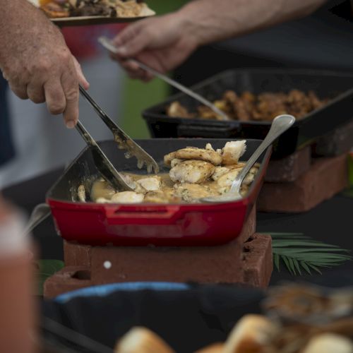 The image shows people serving food from dishes on a table, with utensils and condiments nearby, likely at a buffet or gathering.