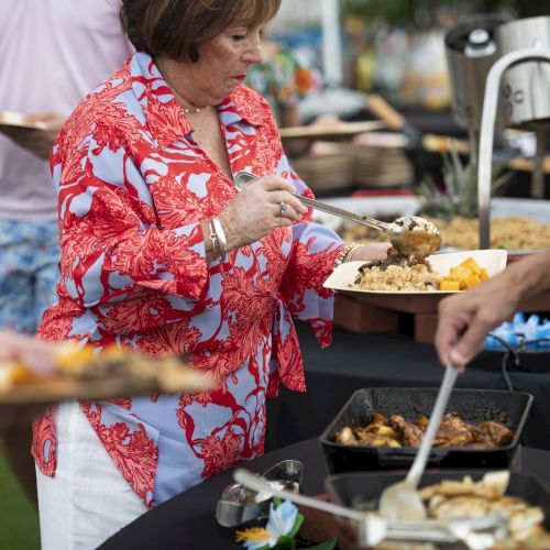A person in a red and white shirt is serving food at a buffet with dishes on a black tablecloth outdoors.