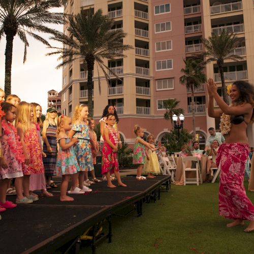 A group of children watches a woman doing a hula dance outside near a tall building with palm trees.