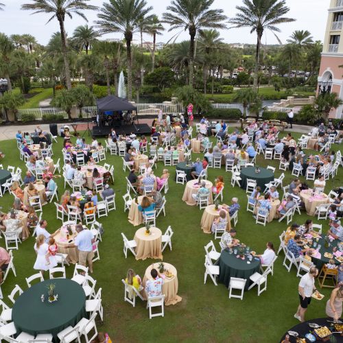 A large outdoor gathering with people seated at tables on a lawn surrounded by palm trees and buildings, creating a festive atmosphere.