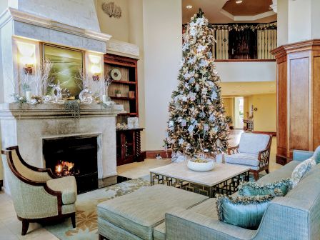 Cozy living room with a tall decorated Christmas tree, fireplace, and soft teal seating arranged around a glass coffee table. Stylish, festive warmth.