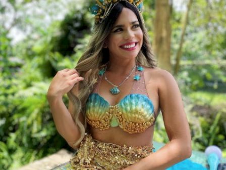 A smiling person dressed as a mermaid with a colorful outfit and headpiece, sitting in a lush outdoor setting with trees in the background.