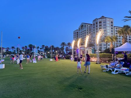 A group of people enjoy an outdoor event on a grassy field with fireworks, tents, and a building in the background at twilight.