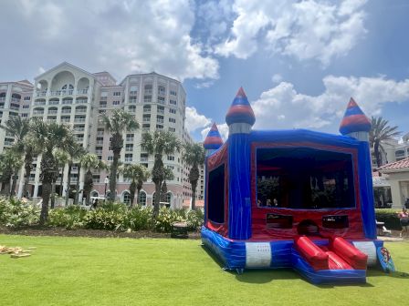 A colorful inflatable bounce house on a grassy area with palm trees and a tall building in the background.