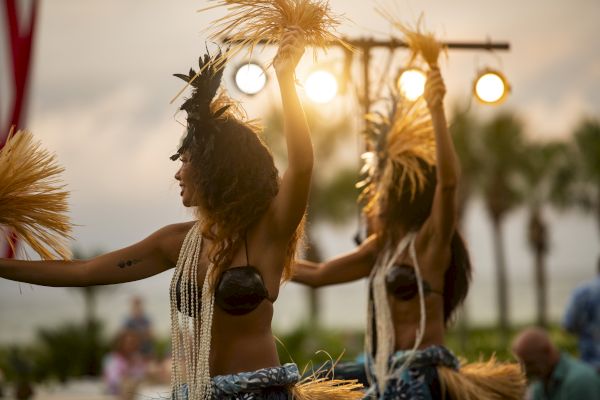 Two dancers perform with traditional attire and accessories at an outdoor event, with palm trees and lights in the background.