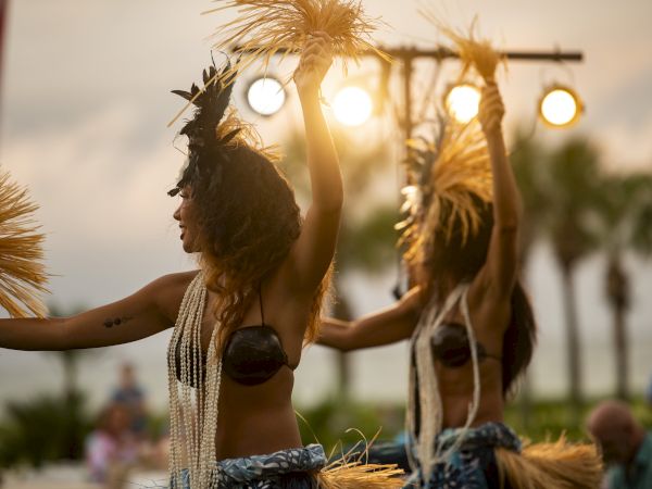 Two dancers perform with traditional attire and accessories at an outdoor event, with palm trees and lights in the background.