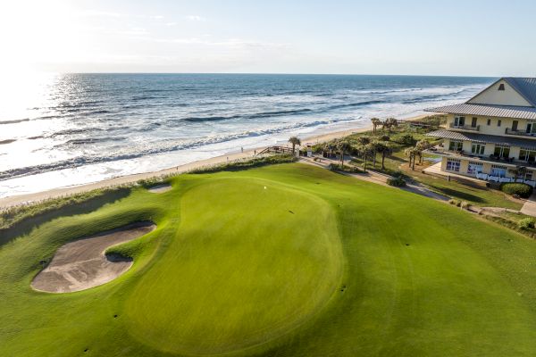 A coastal golf course with a manicured green, a sand trap, and a building nearby, overlooking the ocean under a clear sky.