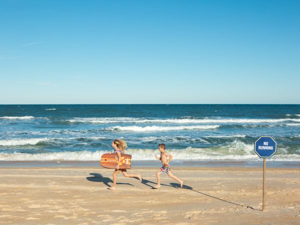 Two children are joyfully running on a sandy beach near the ocean, next to a sign that says "No Running."