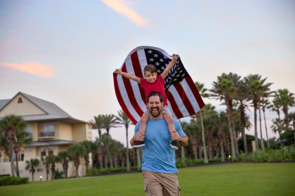 A man carries a child on his shoulders holding a U.S. flag. They are outdoors with palm trees and a building in the background, smiling joyfully.