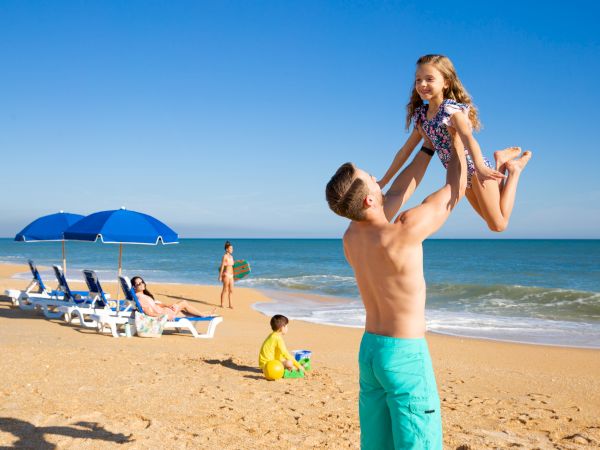 A man lifts a child joyfully on a sunny beach, with umbrellas and people relaxing near the shoreline.