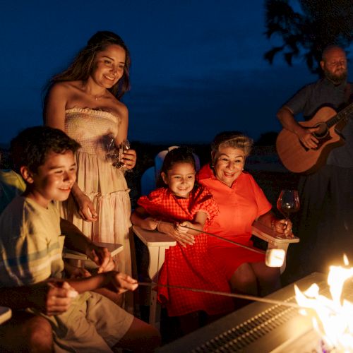 A group enjoys an evening by an outdoor fire, toasting marshmallows, with drinks in hand and live guitar music enhancing the mood.