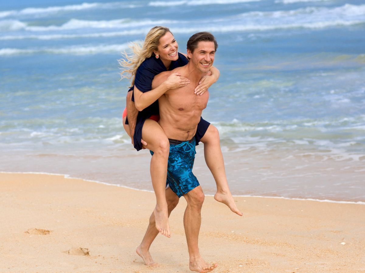 A woman is getting a piggyback ride from a man on a sandy beach by the ocean, both appear happy and relaxed.