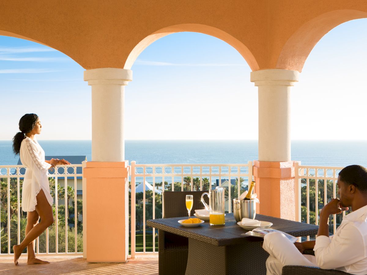 A couple enjoys a seaside view from a terrace, with breakfast on the table under an orange arch, overlooking the ocean under a clear sky.