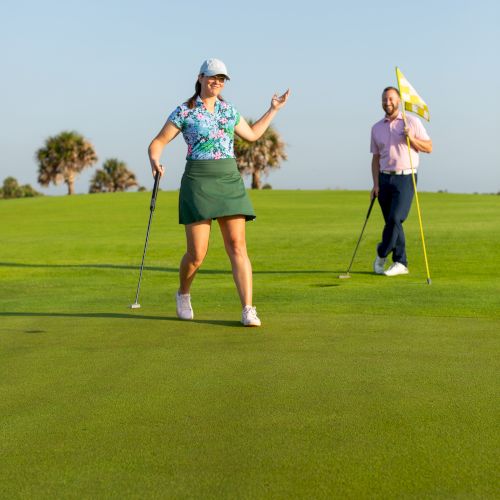 Two people on a golf course, with one holding a flagstick. They appear to be enjoying their game under a clear blue sky.