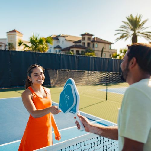 A woman and man are on an outdoor court with paddles, possibly playing pickleball or tennis, with a sunny backdrop.