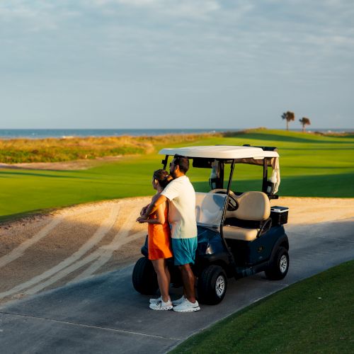 A couple stands near a golf cart on a scenic golf course path, with the ocean and palm trees in the background.