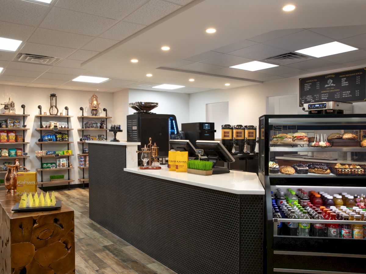 A modern cafe with a counter, coffee machines, snack shelves, and a refrigerated display stocked with drinks and pastries.