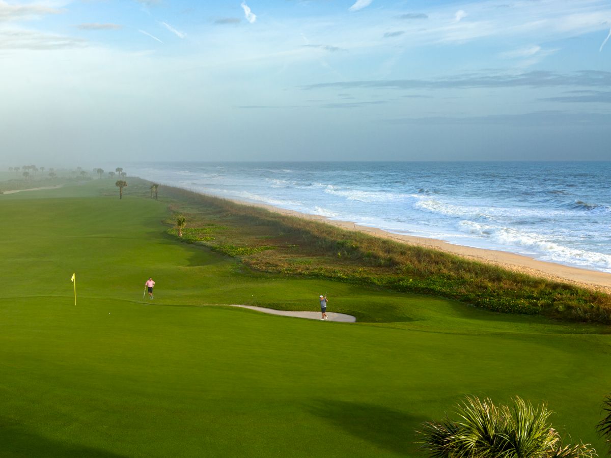 A coastal golf course with two people on the green next to the ocean, featuring bright green grass and waves crashing onto the beach.
