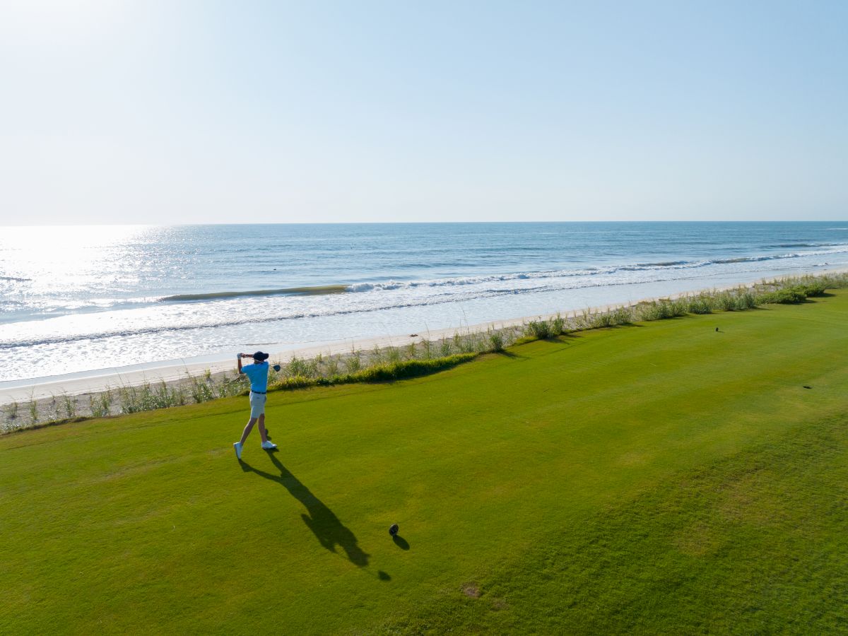 A person is playing golf near a beach, with the sun shining over the ocean in the background.