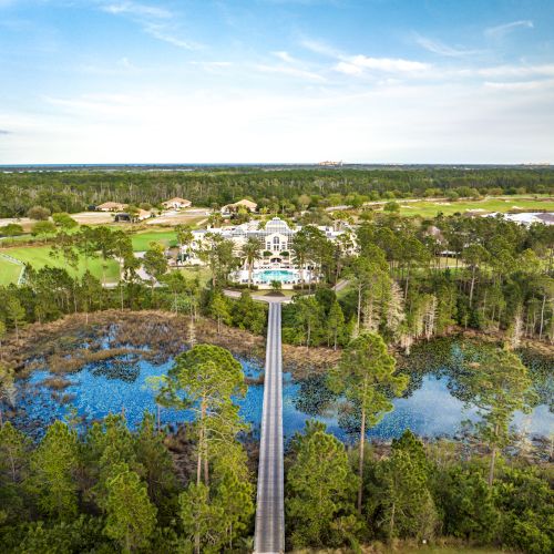 An aerial view of a long pathway over a lake leading to a house surrounded by trees and greenery under a blue sky.