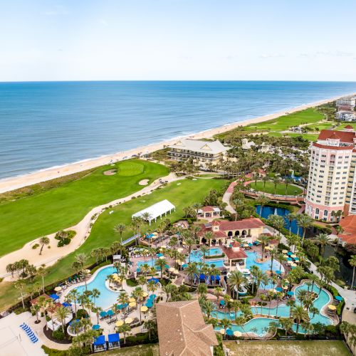Aerial view of a coastal resort with pools, greenery, and beachfront, alongside a golf course and hotels ending at the horizon.