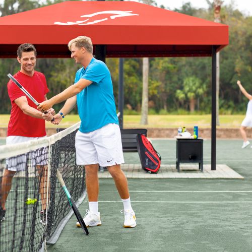 Two people at a tennis court are shaking hands over the net, with others playing in the background near a red canopy.
