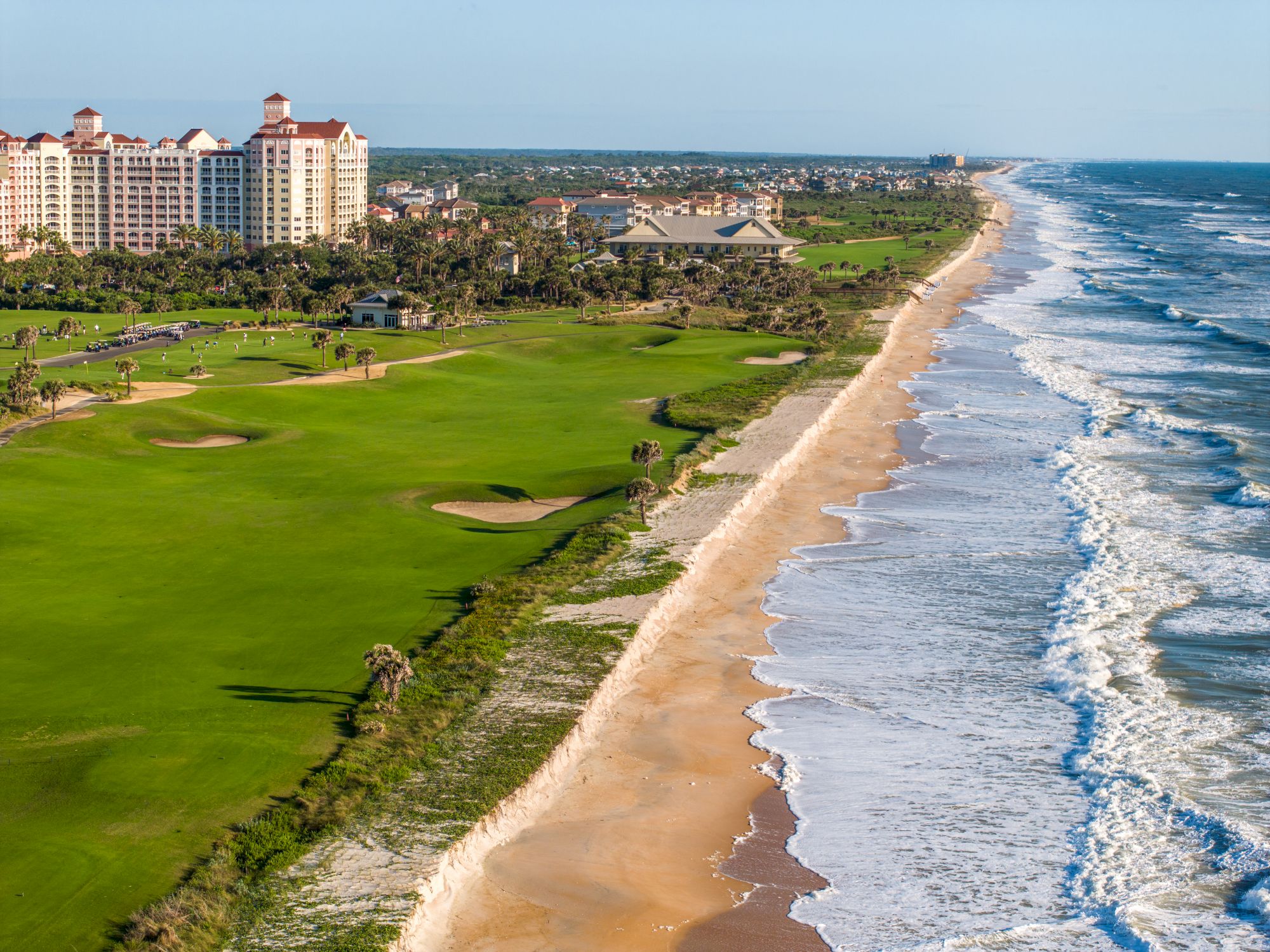 A beachfront landscape with a golf course, sandy shore, ocean waves, and buildings in the background.