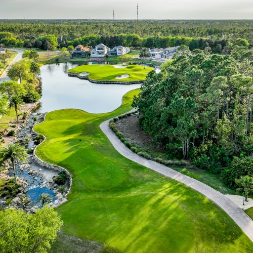 An aerial view of a golf course with a pond, winding paths, and surrounding greenery. Houses are in the background, bordered by forest.