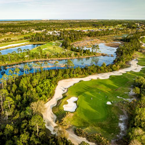 Aerial view of a lush golf course surrounded by water and trees, featuring fairways, sand traps, and a scenic landscape under a clear sky.