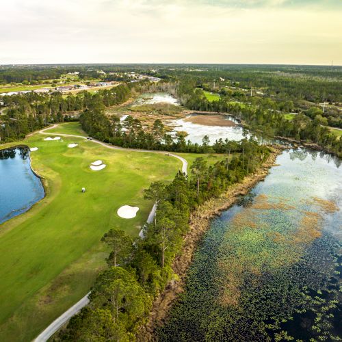An aerial view of a golf course with lush greenery, water features, trees, and sand traps, set in a vast natural landscape.
