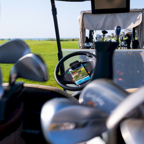The image shows golf clubs in a cart on a golf course, with another cart visible in the background, set against a clear sky.