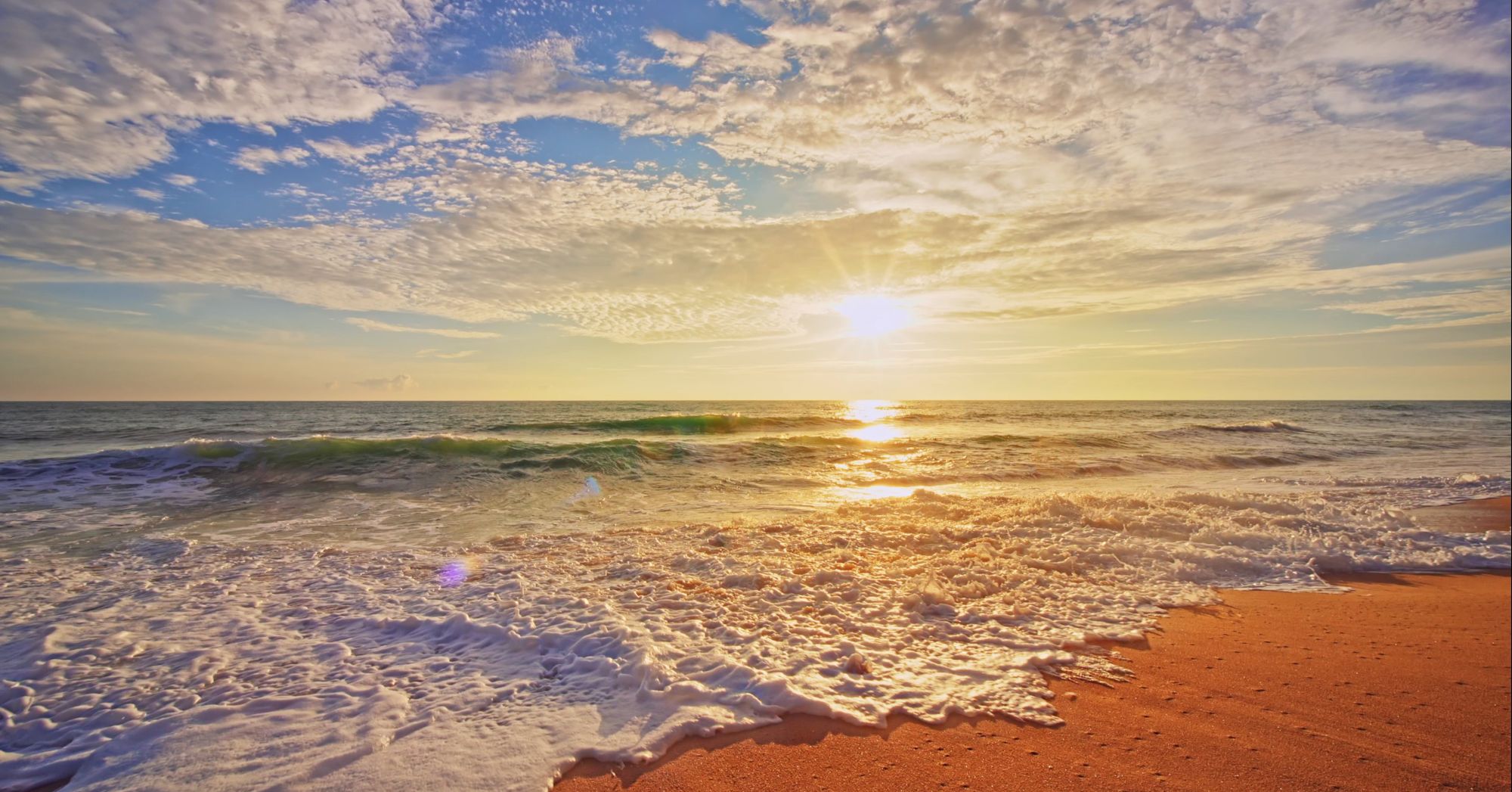 A sunny beach with golden sand, gentle waves, and a bright sun low on the horizon, casting warm reflections across the water.