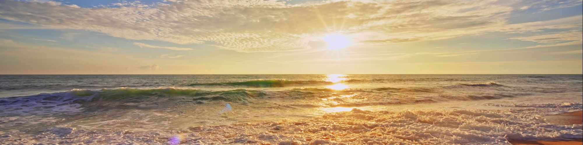 A sunny beach with golden sand, gentle waves, and a bright sun low on the horizon, casting warm reflections across the water.
