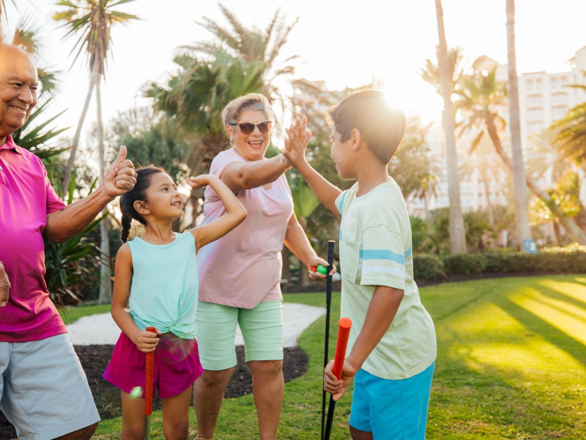 A group of people, including children and adults, enjoying mini-golf outdoors, with trees and sunlight in the background.
