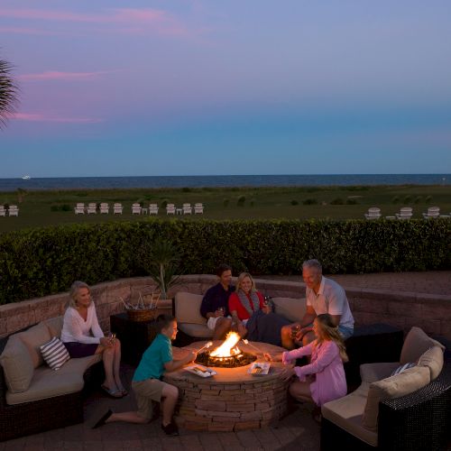 A group of people sits around a fire pit on a patio overlooking a grassy field and ocean at sunset, enjoying a relaxing evening together.
