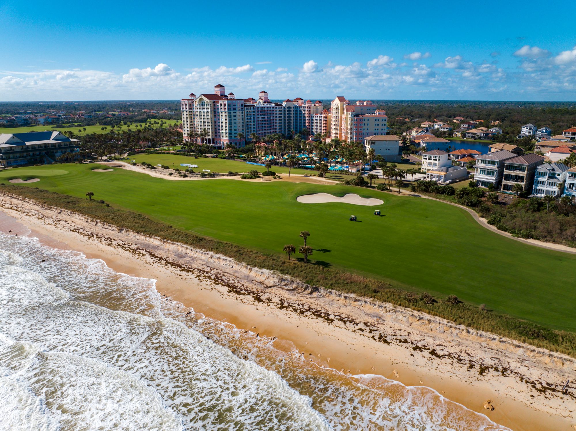 A coastal resort with a golf course, buildings, and a sandy beach alongside ocean waves under a clear blue sky.