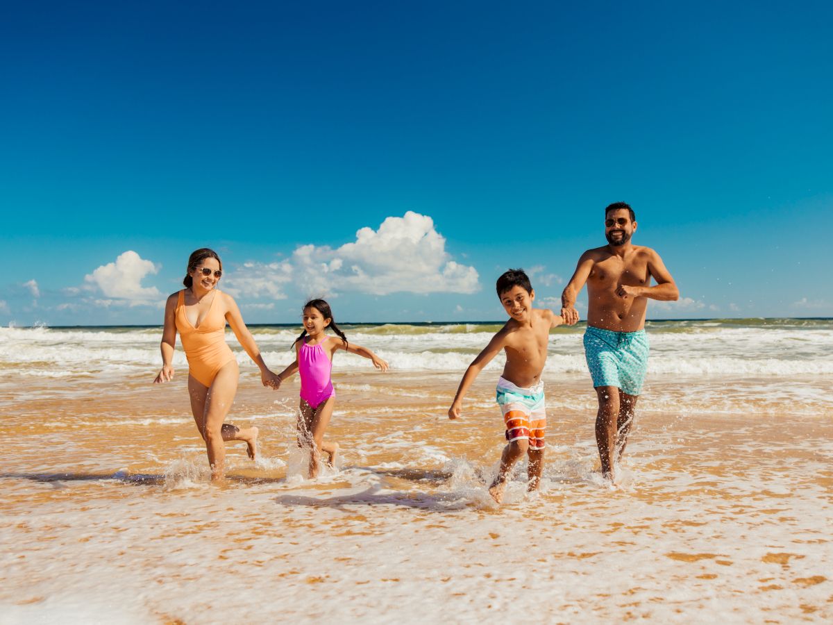 A family of four, two adults and two children, joyfully running through shallow ocean waves on a sunny day at the beach.