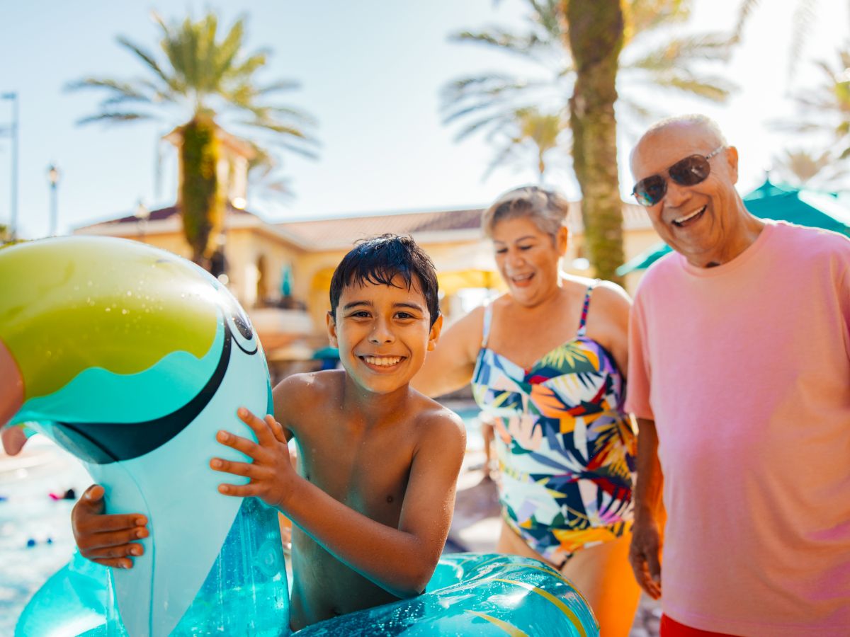 A boy with a pool float is smiling with two adults by a pool. They're all enjoying a sunny day, with palm trees in the background.