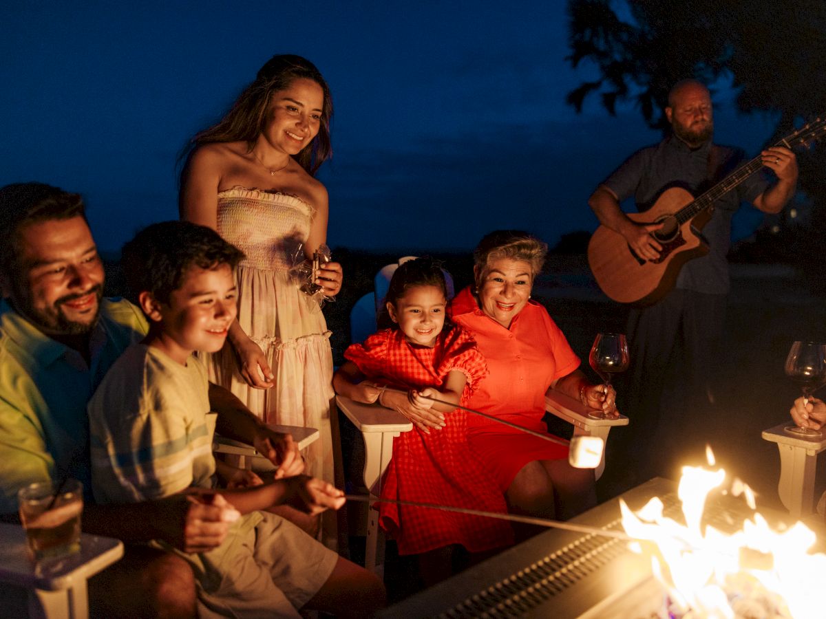 A group of people gathered around a fire, roasting marshmallows, playing guitar, and enjoying a lively evening outdoors.