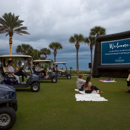 People are enjoying an outdoor movie night with golf carts, a large screen, and a snack stand under palm trees.