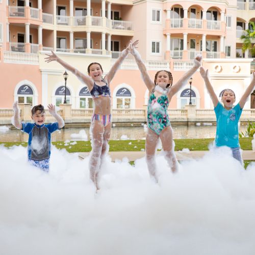 Children joyfully play in foam outside a building, with adults watching, creating a lively and fun atmosphere.