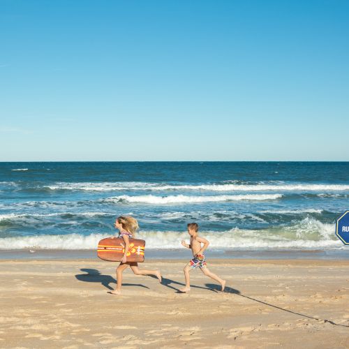 Two children running on a beach near the water's edge, carrying a bodyboard; a "No Running" sign is visible in the sand.